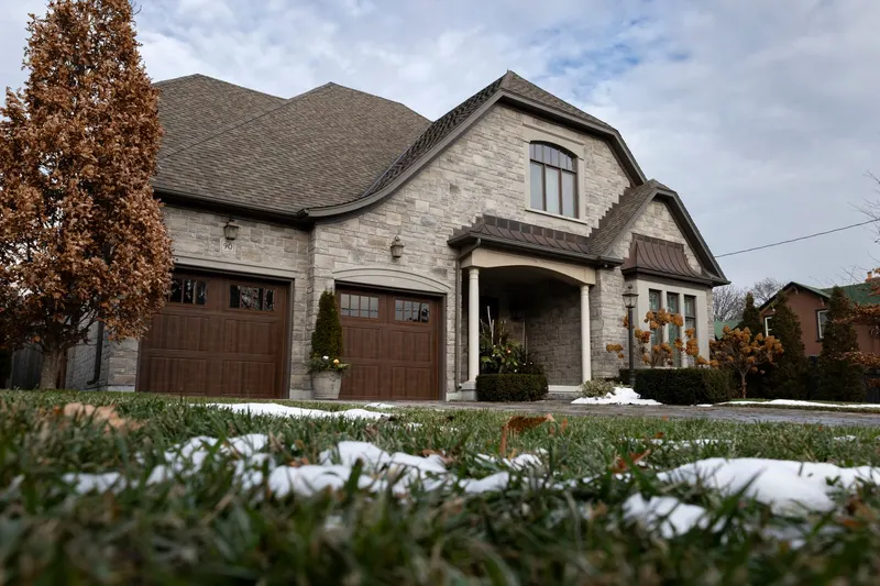 Two brown carriage-style garage doors on a stone house, with patches of early snow on the front lawn