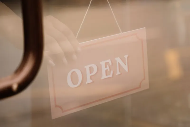 A hand turning a hanging Open sign on the glass door of a shop, next to a curved metal door pull