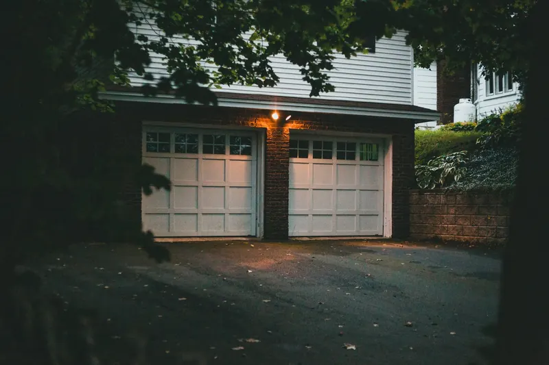 A brick double garage with two white panel doors and a lit wall lamp, seen from a dark driveway at dusk