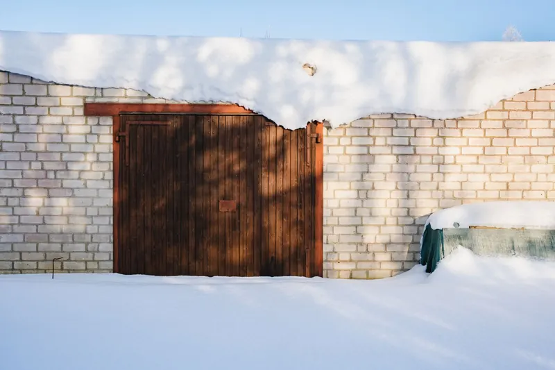 A wooden garage door set in a white brick wall, with deep snow drifted against its base