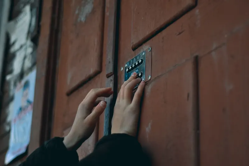 Two hands in dark sleeves pressing the buttons of a keypad lock on a weathered red door