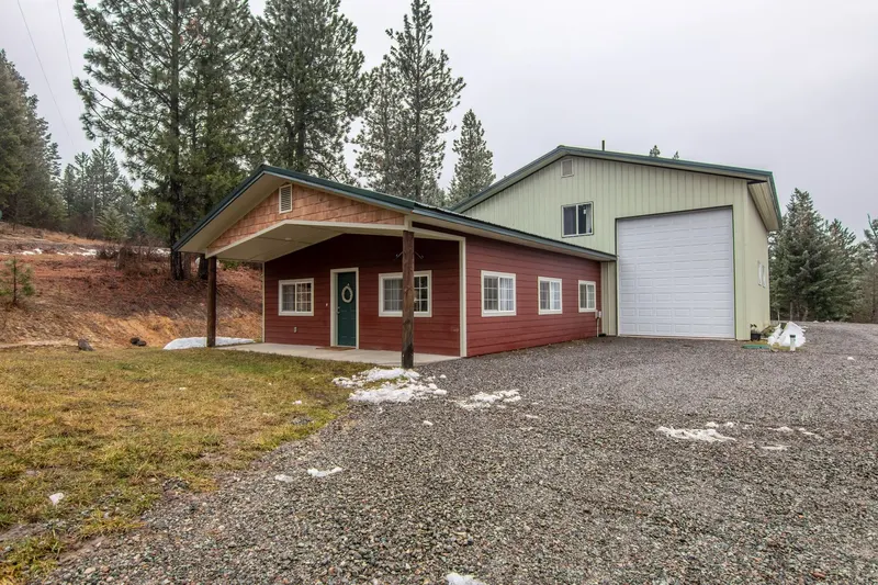 A red house with a tall attached shop garage and a white overhead door, with patches of snow on the gravel drive