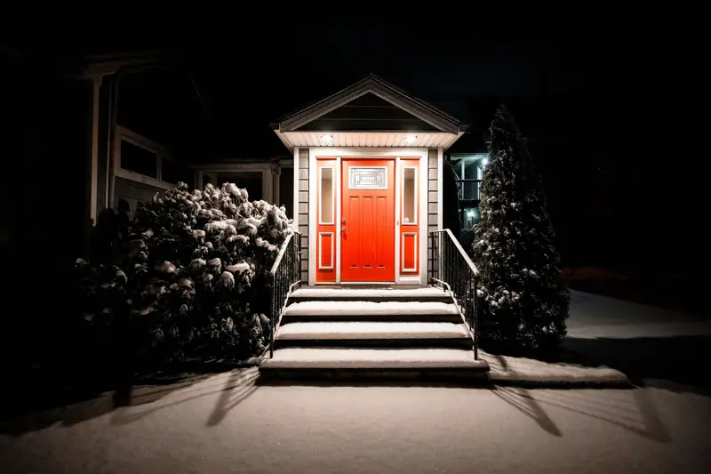 A front porch at night in fresh snow, the porch light on above a red front door