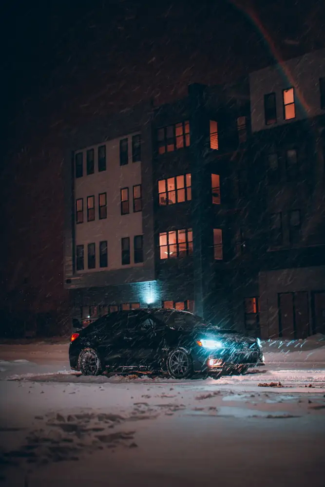 A car with its headlights on in a snowy parking lot at night