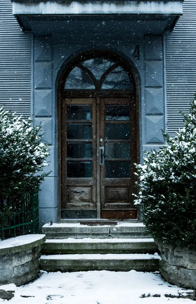 An old wooden front door above snowy steps as snow falls